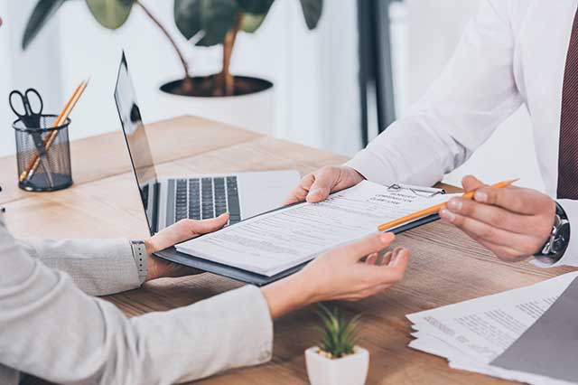 man in white business shirt and tie handing documents to a woman across a table with a laptop on it